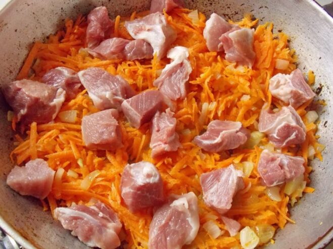 Browning the meat with onions and carrots to lock in flavor. Pieces of raw meat being added to the skillet with sautéed onions and carrots.