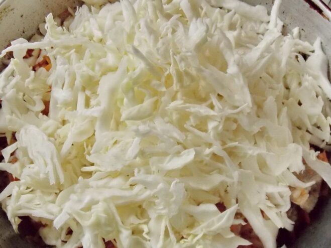 Adding fresh shredded cabbage to the skillet. A large amount of shredded white cabbage being added to the skillet with meat and vegetables.