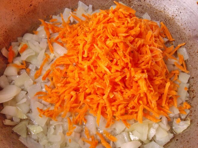 Adding Carrots for Sweetness and Color Grated carrots being added to the skillet with sautéed onions.