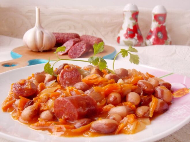 A white bowl filled with stewed beans and smoked sausages in a rich tomato sauce, garnished with fresh parsley.