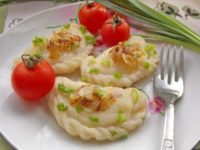 A white bowl filled with freshly boiled potato vareniki, garnished with fried onions and fresh dill.