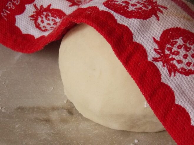 Allowing the vareniki dough to rest under a towel to become more pliable. A ball of vareniki dough resting in a bowl, covered with a kitchen towel.
