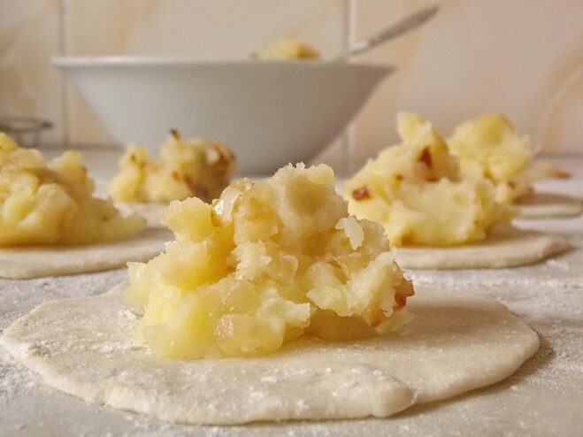 Placing a spoonful of potato filling onto each rolled-out dough circle. Small circles of dough with a spoonful of potato filling on each.