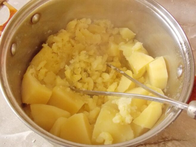 Mashing the boiled potatoes to create a smooth puree for the filling. Boiled potatoes being mashed in a pot to make the filling.