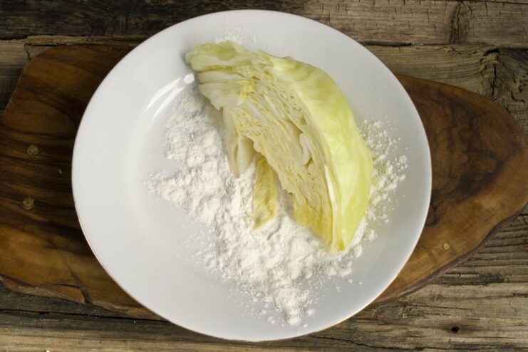 First Breading Step: Flour Coating. This helps the egg wash and breadcrumbs adhere better. A cooked cabbage wedge being coated in a shallow dish of white flour.