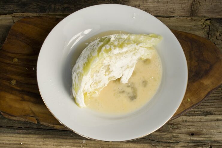 Second Breading Step: Egg Wash. Ensure the entire wedge is evenly coated in the beaten egg. A flour-dusted cabbage wedge being dipped into a bowl of beaten eggs.