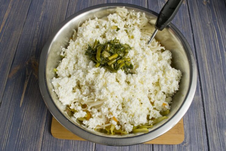 Adding Creaminess: Mixing in Sieved Tvorog and Greens Sieved tvorog and chopped greens being added to the pasta and vegetable mixture in a bowl.