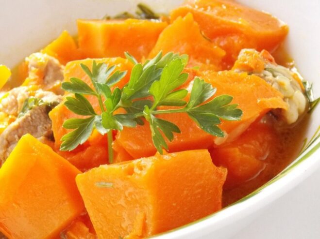A white bowl filled with vibrant orange pumpkin and pork stew, garnished with fresh green herbs.