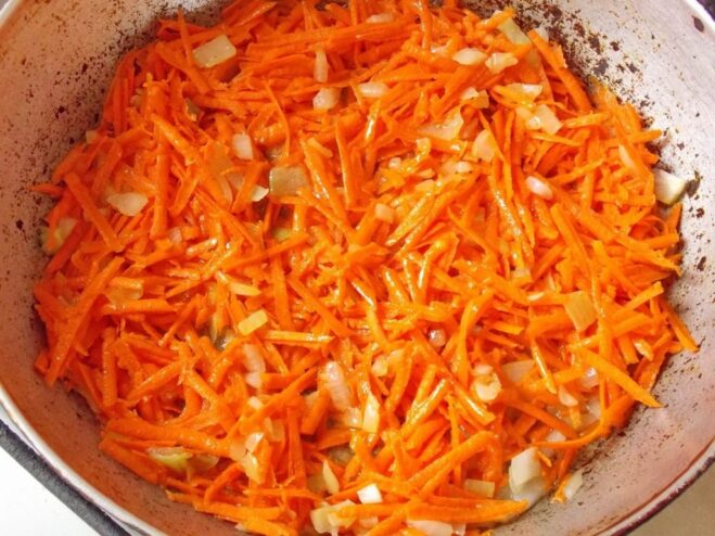 Adding grated carrots to the sautéed onions Grated carrots being added to the pan with translucent onions.