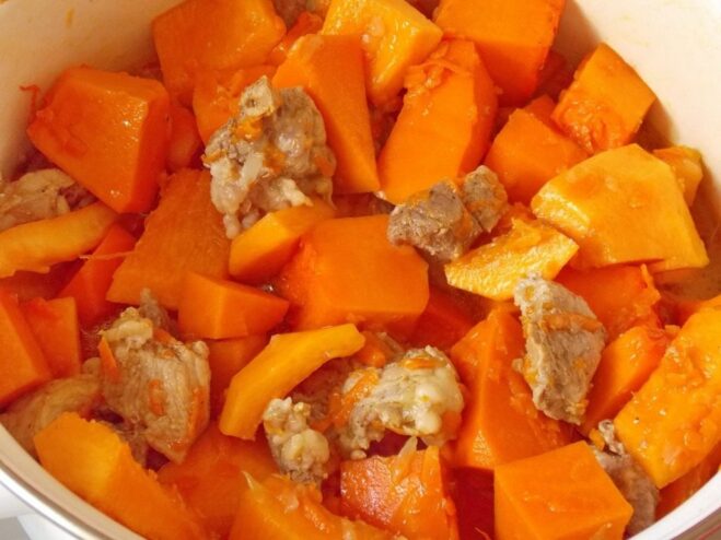 Adding large chunks of pumpkin to the stew Large cubes of bright orange pumpkin being added to the pot with the simmering meat.