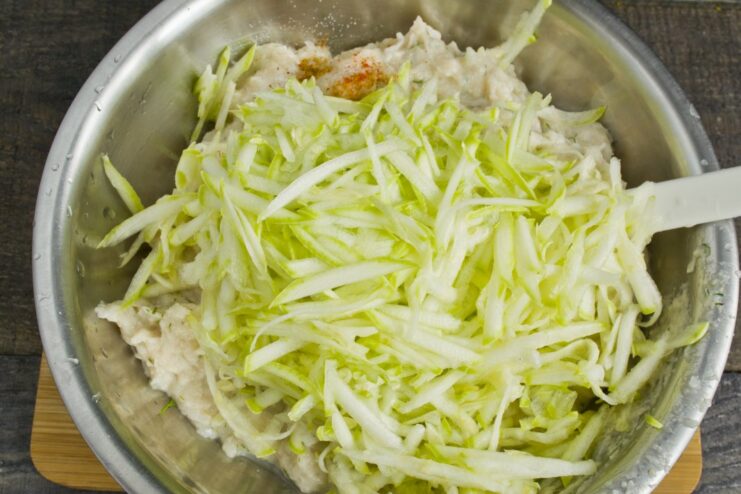Grating fresh zucchini to add moisture and flavor to the chicken patties. A hand grating a fresh green zucchini on a box grater.