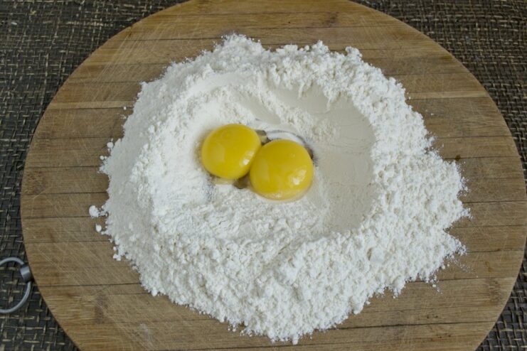 Step-by-Step: Making Fresh Pasta Dough for Lasagna Hands kneading a ball of fresh pasta dough on a wooden board with flour and eggs nearby.