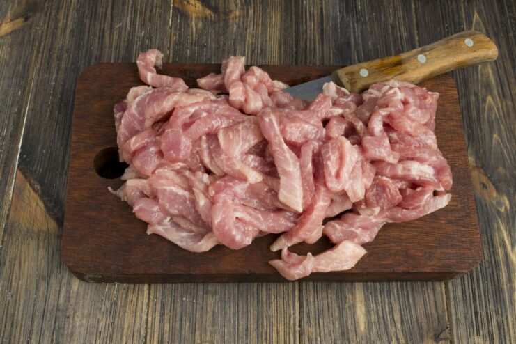 Adding strips of lean pork to the pan with sautéed onion and garlic Strips of lean pork being fried with onions and garlic in a pan.