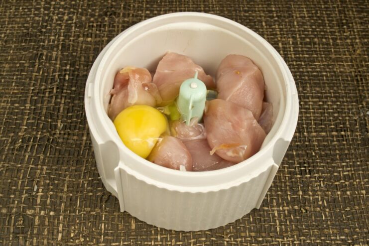 Adding an egg to bind the chicken filling A raw egg being added to chopped chicken fillet in a blender bowl.