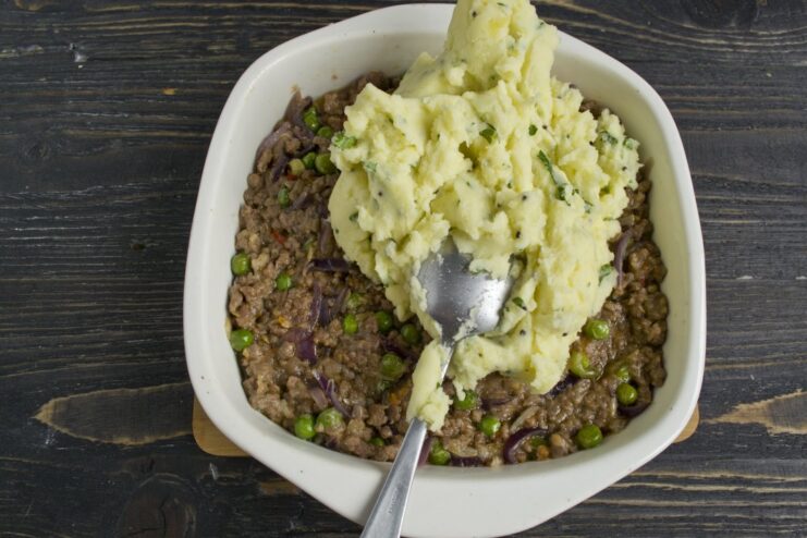 Assembling the pie: layering the savory meat filling in a baking dish, ready for the potato topping. A baking dish with a layer of minced meat filling, with mashed potatoes being added on top.
