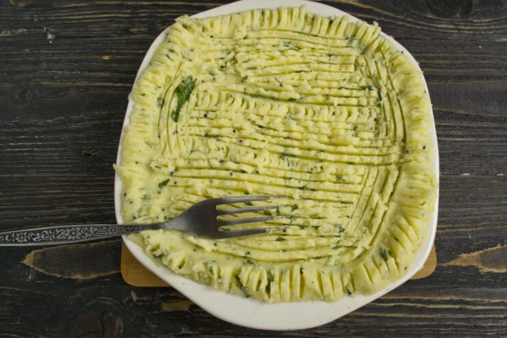 Creating a decorative, wavy texture on the potato topping with a fork to ensure a crispy crust. Mashed potato topping being spread over the pie filling, with fork marks creating a pattern.