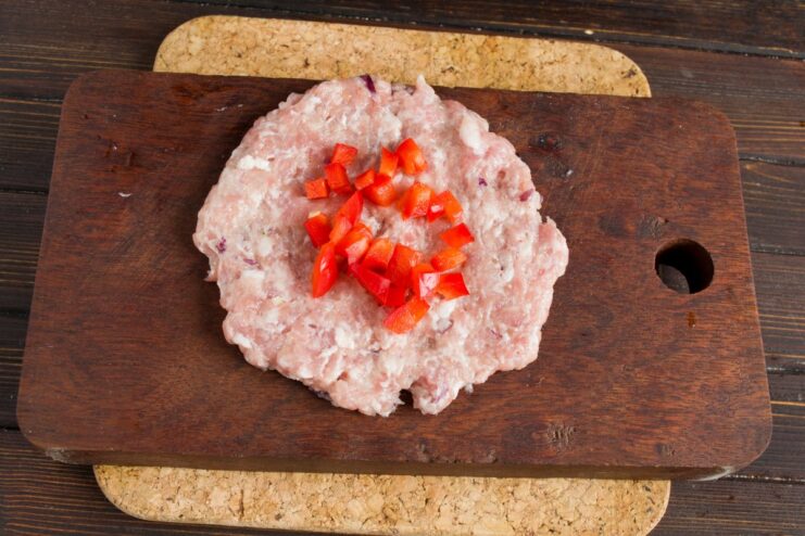 Adding the Sweet Pepper Filling to the Cutlet Chopped red bell pepper being placed in the center of a raw meat patty.