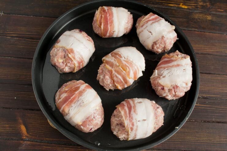 Arranging Bacon-Wrapped Cutlets for Baking Several bacon-wrapped stuffed cutlets arranged in an oven-safe skillet, ready for baking.