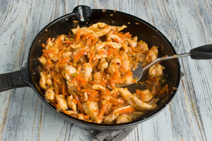Combining Chicken and Sauce for the Final Step Cooked chicken pieces being mixed with vegetables in a thick sweet and sour sauce in a pan.