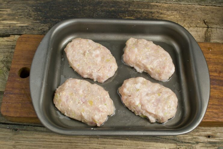 Shape the mixture into oval patties before baking. Raw meat patties neatly arranged on an oiled baking sheet, ready for the oven.