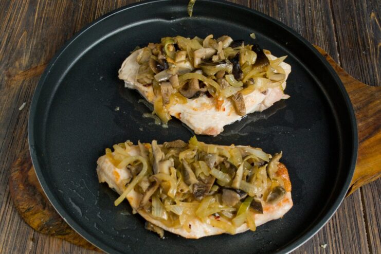 Cooked mushroom and onion mixture being spread over seared chicken cutlets on a baking sheet.