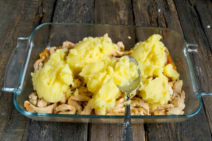 Spoonfuls of mashed potatoes being added on top of the chicken filling in a baking dish.