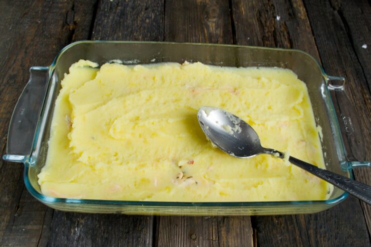 A spatula spreading mashed potatoes evenly over the filling in a baking dish.