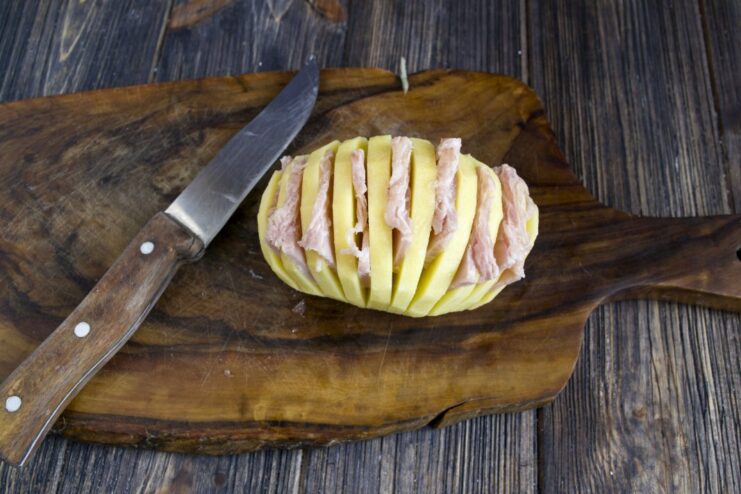 Step 3: Stuffing the Potato with Ham A raw accordion potato with thin slices of ham inserted into each cut.