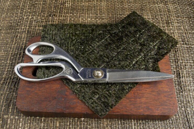 Preparing Nori Sheets for Temaki by Cutting Them in Half A stack of nori seaweed sheets being cut in half with a knife.