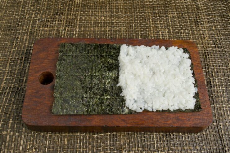 Spreading a Layer of Sushi Rice onto a Nori Sheet A hand spreading sushi rice over half of a nori seaweed sheet.