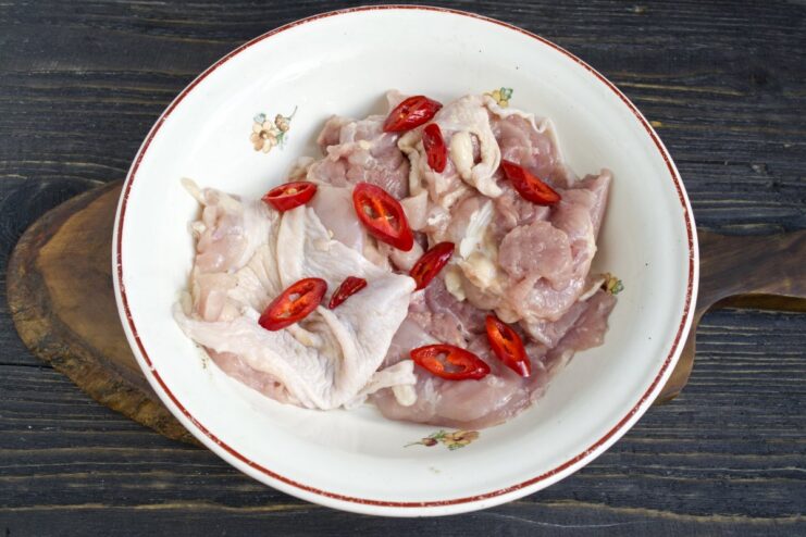 Adding Peppers to Chicken for Extra Flavor Deboned chicken thighs in a bowl with sliced red chili peppers being added.