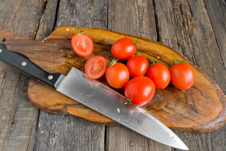 Halving cherry tomatoes to release their juices while roasting. Red cherry tomatoes being sliced in half on a cutting board.