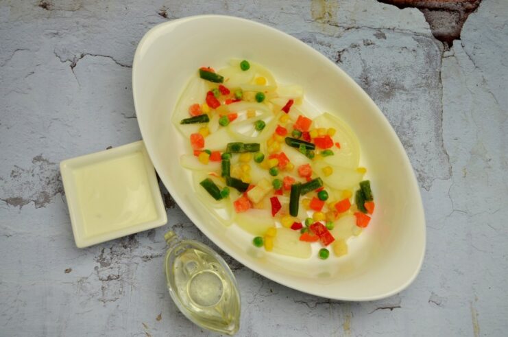 A layer of frozen mixed vegetables over sliced onions in a baking dish, drizzled with oil.