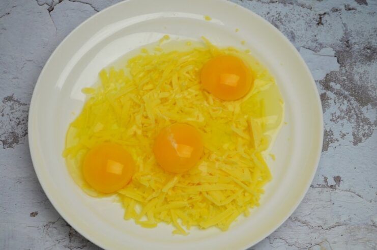 Three cracked eggs in a bowl with grated cheese, ready to be mixed.