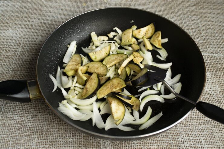 Adding eggplant slices to the vegetable mix Thinly sliced eggplant being added to the pan with other vegetables.