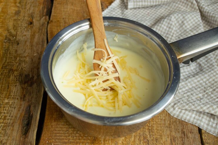 Grated cheese melting into the cream sauce in a saucepan.