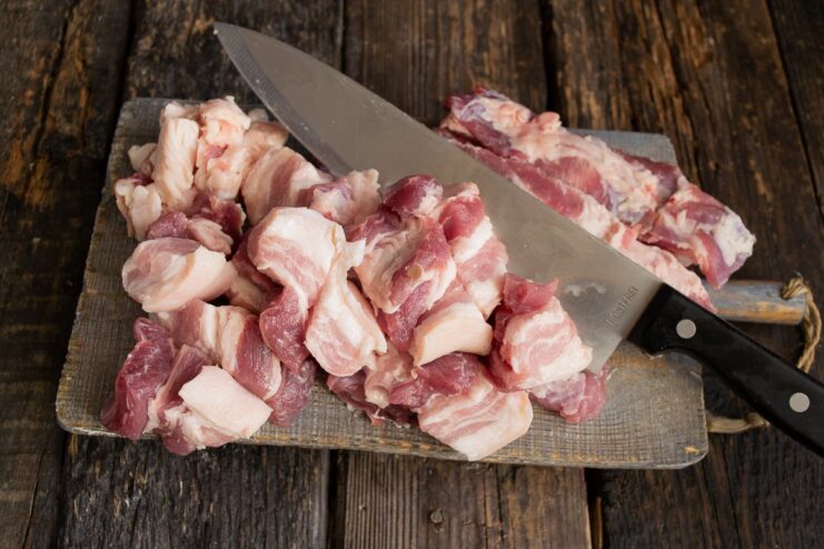 Slicing fresh pork belly into chunks for the Bigos stew. Raw pork belly being cut into large chunks on a cutting board.