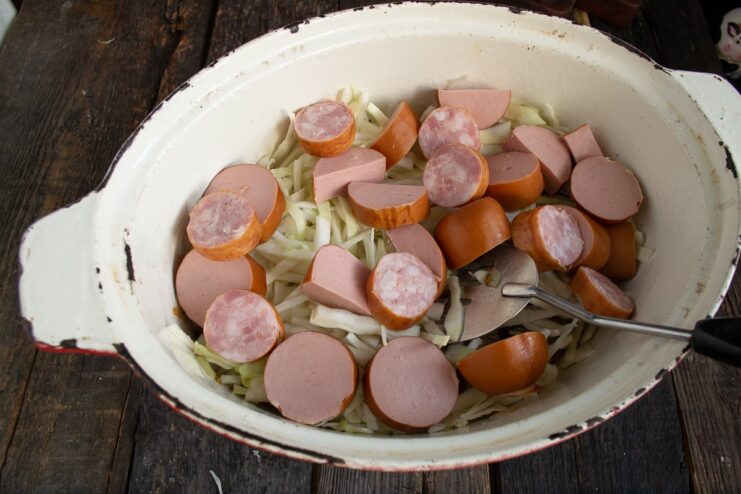 Adding smoked sausages and sardelki to the Bigos. Sliced smoked sausage and sardelki being layered over fresh cabbage in the pot.