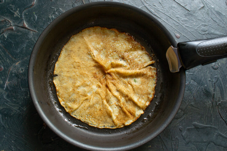 How to fry the perfect golden-brown crepe in a hot pan. A thin crepe being fried in a black skillet until golden brown.