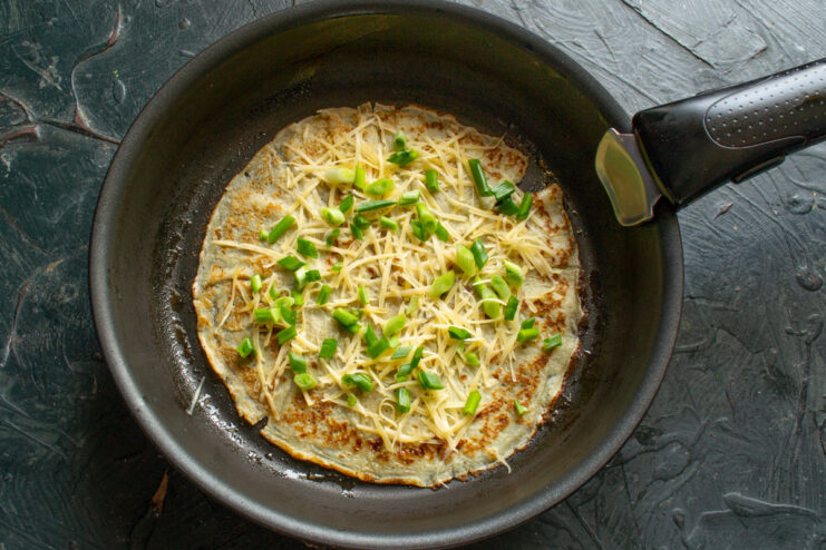 Adding the cheese and green onion filling to the crepe. A crepe in a pan being topped with grated cheese and chopped green onions.