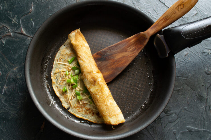 How to roll a filled crepe neatly in the pan. A filled crepe being rolled into a tube shape with a spatula in a pan.