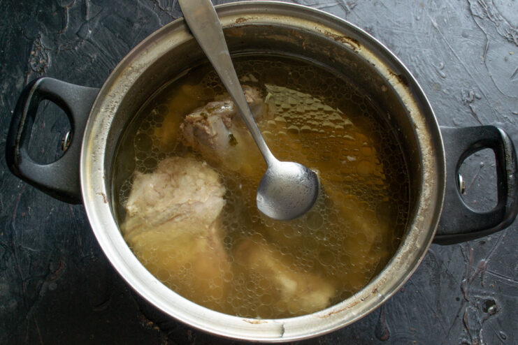 A pot of borscht broth with ribs simmering on the stove, with foam being skimmed off.