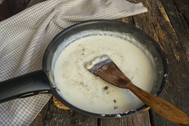 Creating a smooth and creamy sauce by whisking in cream and seasonings. Cream being whisked into a roux in a skillet to create a smooth white sauce.