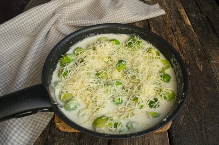 Topping the Brussels sprouts with Parmesan cheese before baking. Brussels sprouts in cream sauce in a skillet, being topped with grated Parmesan cheese.