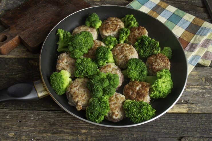 Adding Blanched Broccoli to the Pan with Patties Bright green blanched broccoli florets being arranged in a skillet between pan-fried meat patties.