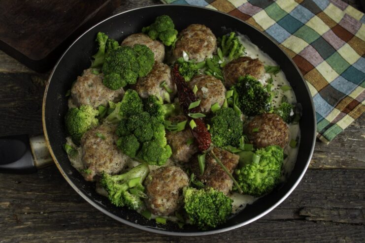 Finishing the Dish with Creamy Béchamel and Fresh Garnish A skillet with meat patties and broccoli, with creamy béchamel sauce being poured over, ready to be served.