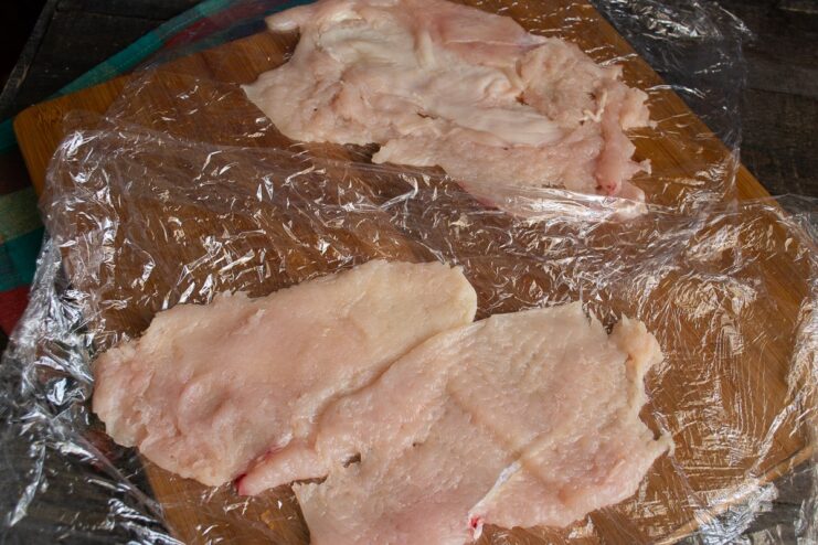 Assembling the Roll's Outer Layer Two thin slices of pounded chicken fillet laid overlapping on plastic wrap.