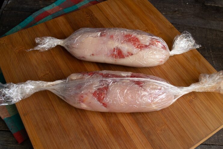 Wrapping the Beef and Chicken Roll The meat roll being tightly wrapped in plastic wrap, with the ends secured.