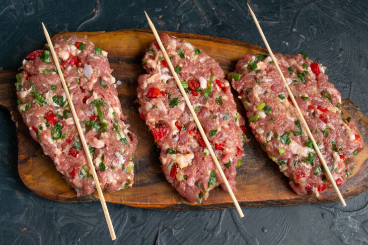 Shaping the Meat Patties Around the Skewers A flat, narrow patty of ground meat with a bamboo skewer placed on top.