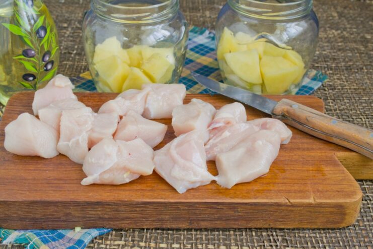 Adding the Protein: Layering Chicken Pieces in the Jars Chunks of raw chicken breast being added on top of the potato layer in glass jars.